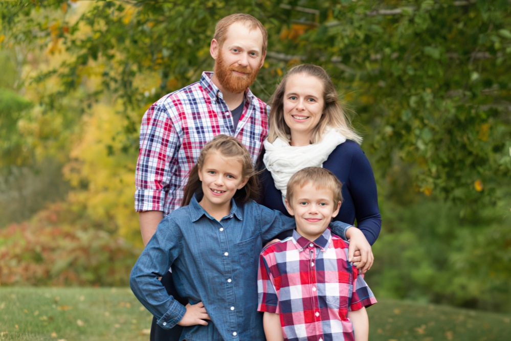 A family smiles together outdoors in matching plaid outfits, surrounded by colorful autumn foliage.