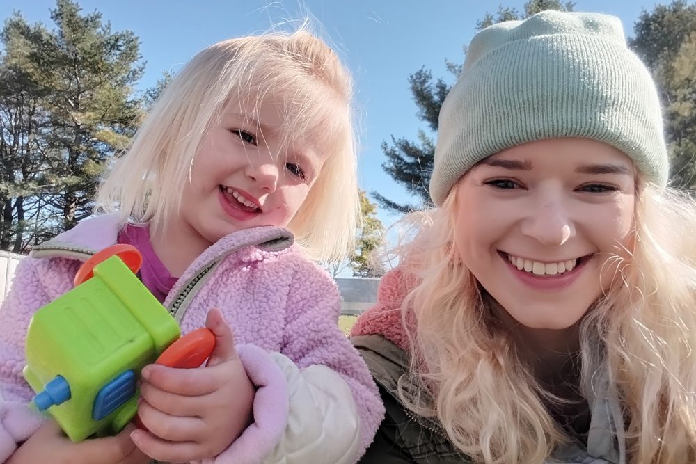 A smiling child holds a toy while posing with her mother, both enjoying a sunny day outdoors.