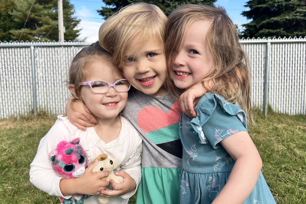 Three cheerful girls hug each other, holding stuffed animals, enjoying a sunny day outdoors.