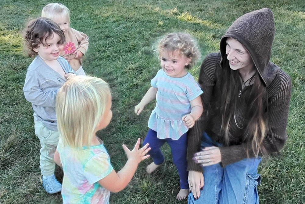 A group of four young children play joyfully in a grassy area, interacting with their teacher, sharing smiles and laughter.