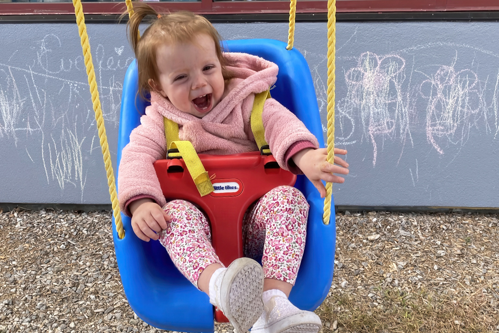 A joyful toddler swings in a bright blue swing, smiling widely, wearing a pink hoodie and colorful leggings.