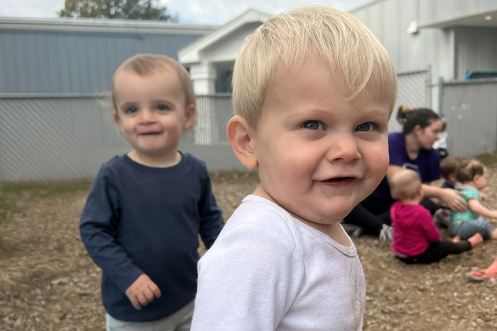 Two smiling toddlers play outdoors, with other children visible in the background, enjoying their time together.