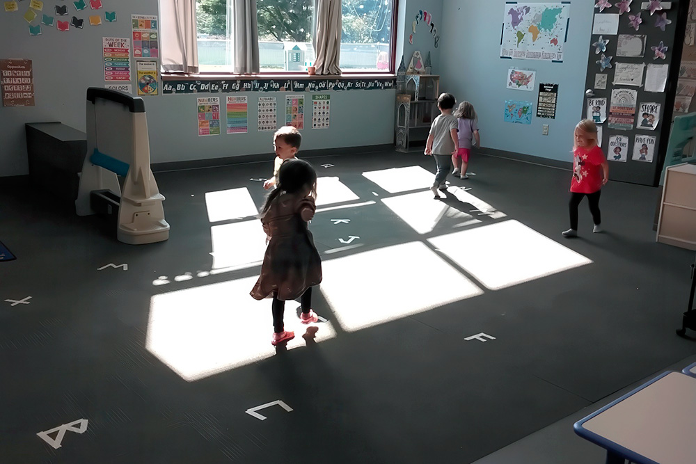 Children play and dance in a brightly lit classroom, with sunlight casting playful shadows on the floor.