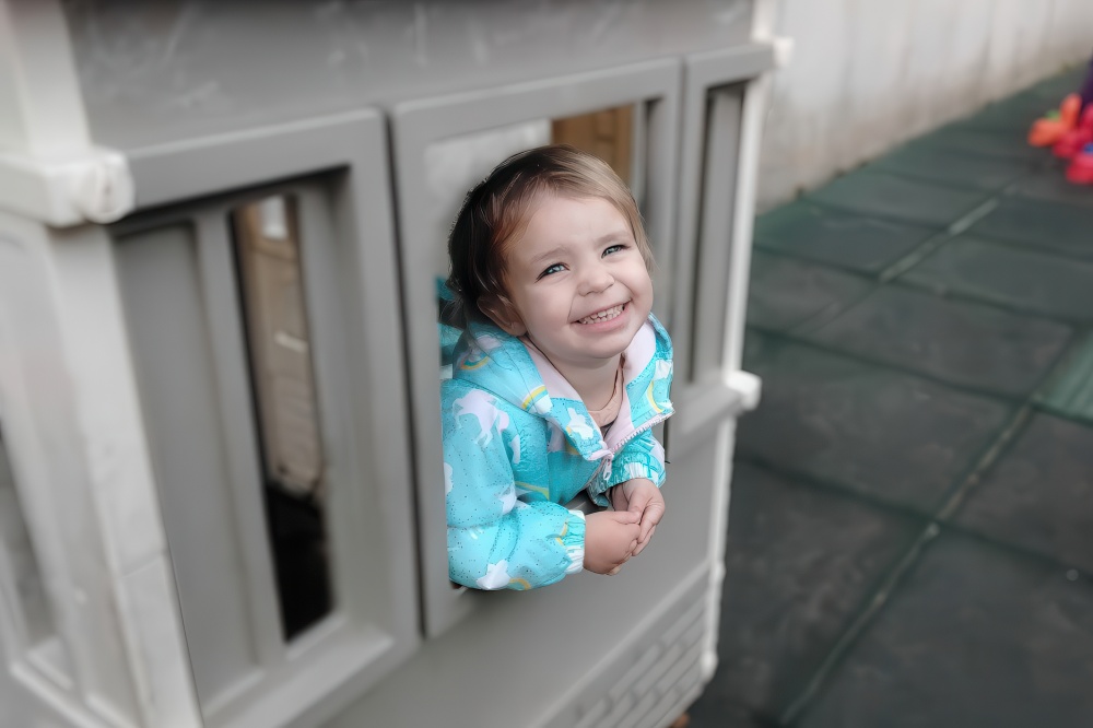 A girl peeks out from a playhouse window, grinning brightly and enjoying her time in the playful setting.