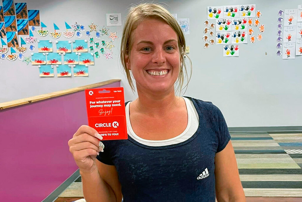 A smiling woman holds a gift card, posing proudly in front of colorful artwork displayed on the wall behind her.