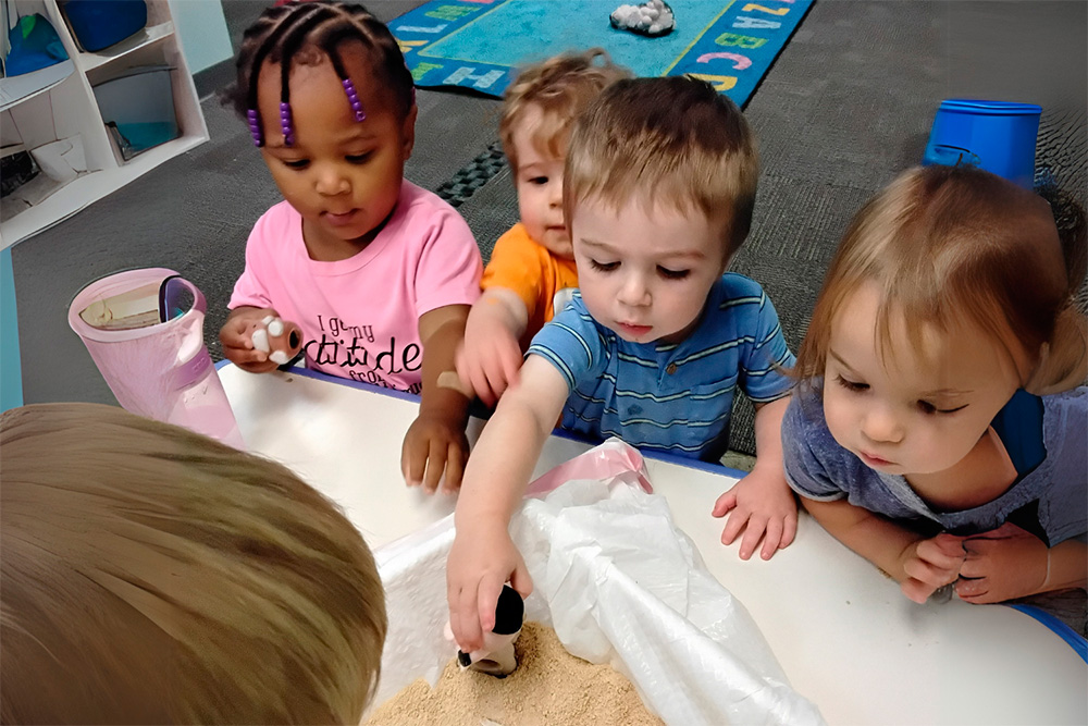 Four children gather around a table, exploring sand with small toys, showing curiosity and engagement in their activity.