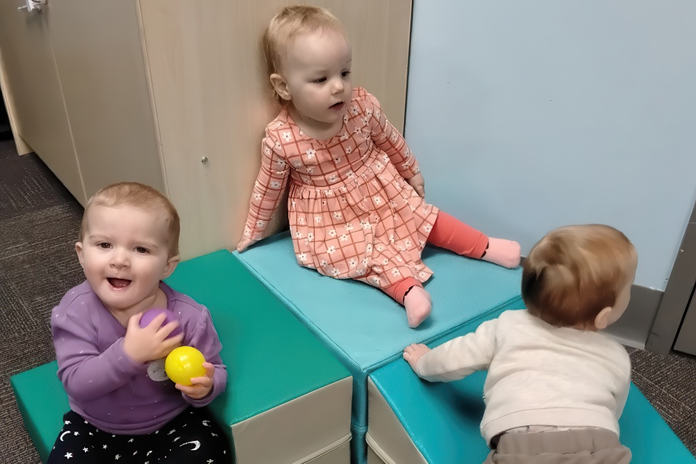 Two toddlers play on colorful soft blocks, one holding a yellow ball and smiling, while another explores nearby.