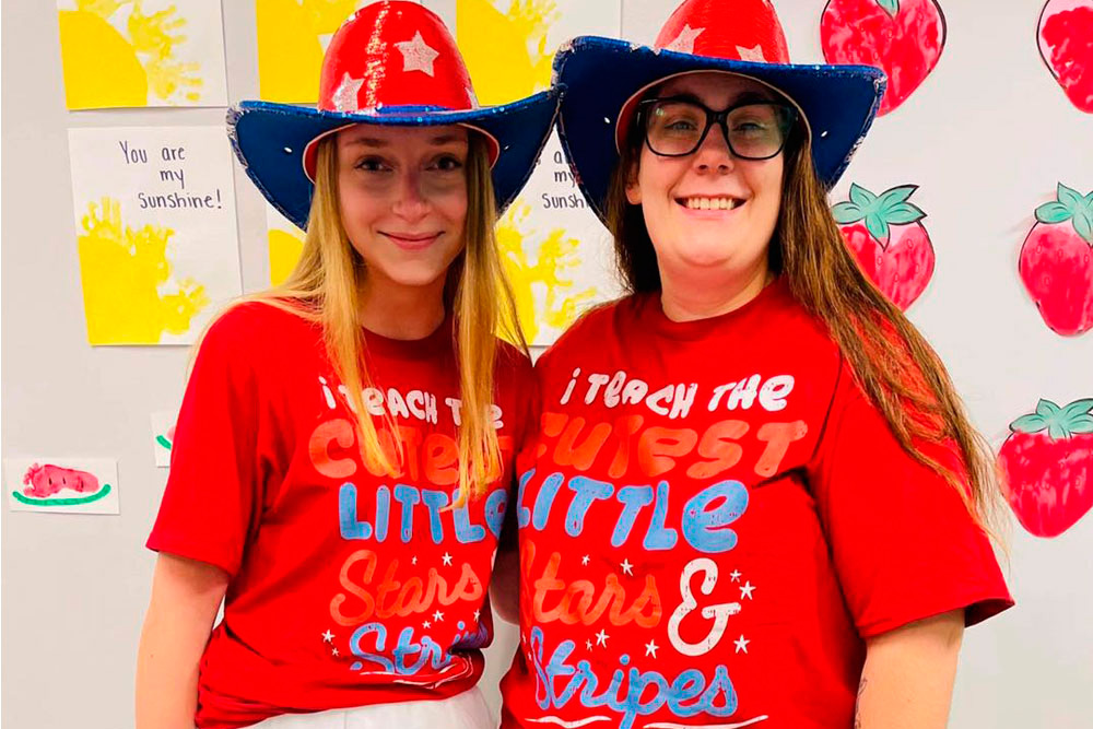 Two women pose happily in matching red shirts and star hats, celebrating their roles as educators with playful spirit.