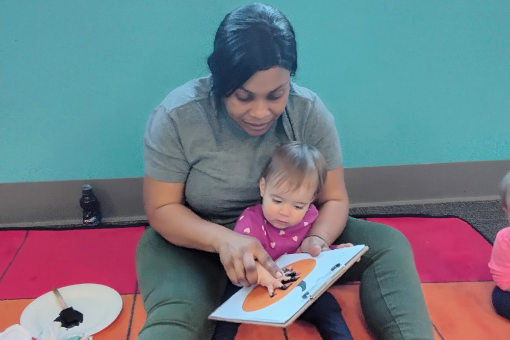 A teacher sits on the floor with a toddler, engaging in an art activity, as they work on a project with paint in clipboard.