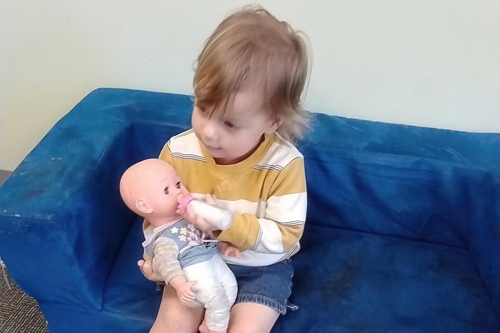 A toddler sits on a blue couch, gently feeding a doll with a bottle, engaged in imaginative play.