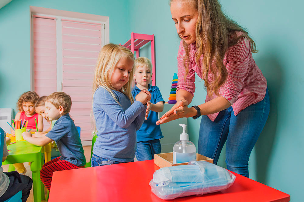 A teacher demonstrates hand sanitizing to a child, while other kids engage in activities at a vibrant, playful setting.