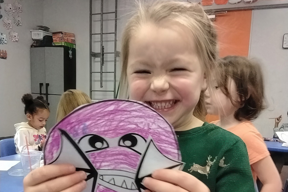 A girl proudly holds up a colorful shark drawing, smiling widely, with other children working on art projects at the back.