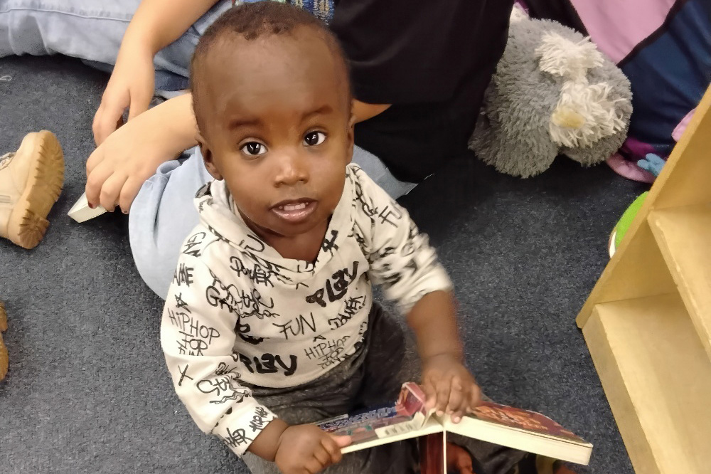 A toddler sits on the floor, smiling at the camera while holding a book, with soft toys visible in the background.