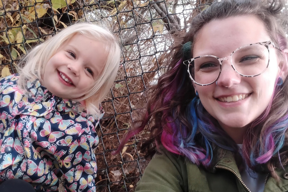 A young girl and a teacher smile together, enjoying a playful moment outdoors, framed by nature and a chain-link fence.