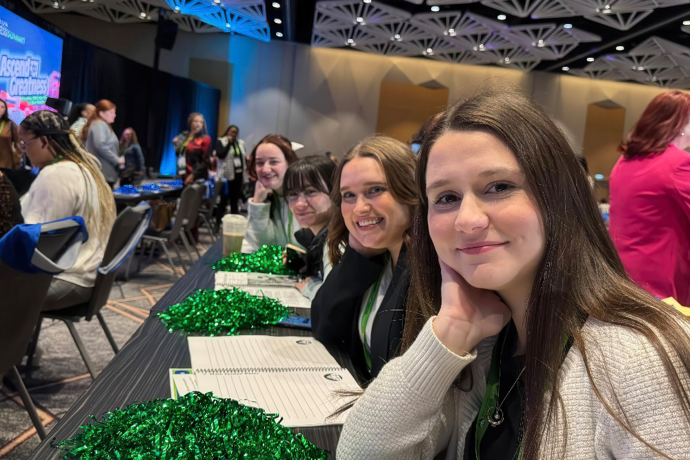 Four young women smile at the camera during a conference, sitting at a table adorned with notebooks and festive decorations.