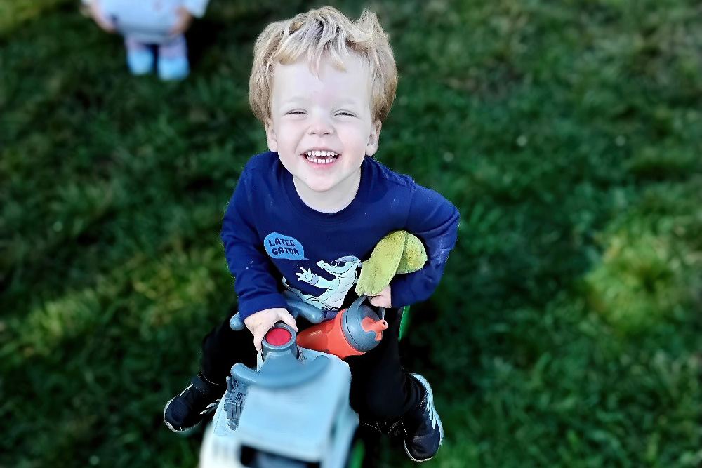 A young boy smiles at the camera while riding a toy outside, holding a stuffed animal, clearly enjoying playtime.