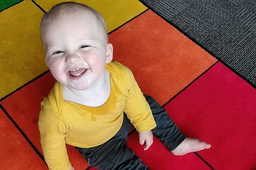 A happy toddler sits on a vibrant, multi-colored mat, grinning widely with a joyful expression.