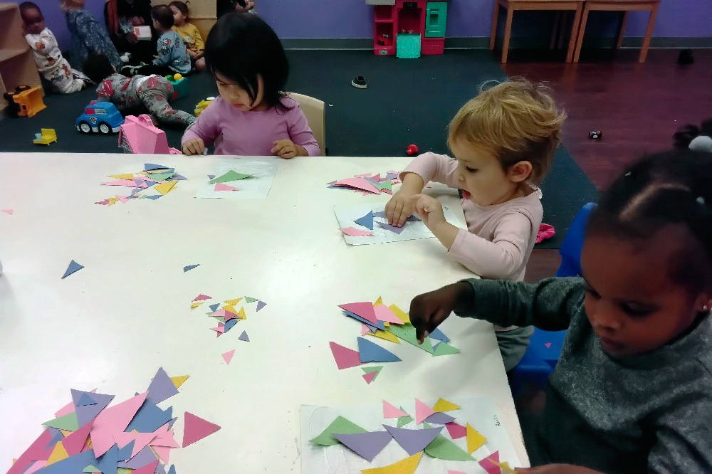 our kids making craft with colorful paper triangles at a table, while another child plays at the back.