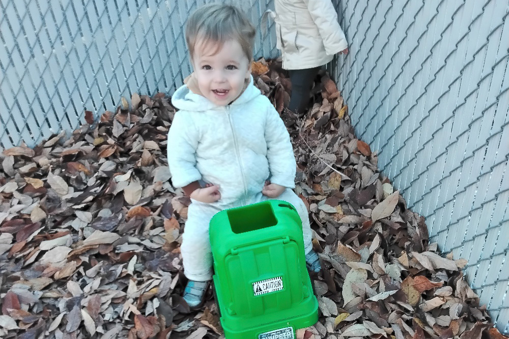 A smiling toddler sits on a green toy in a pile of autumn leaves, enjoying a playful moment outdoors.