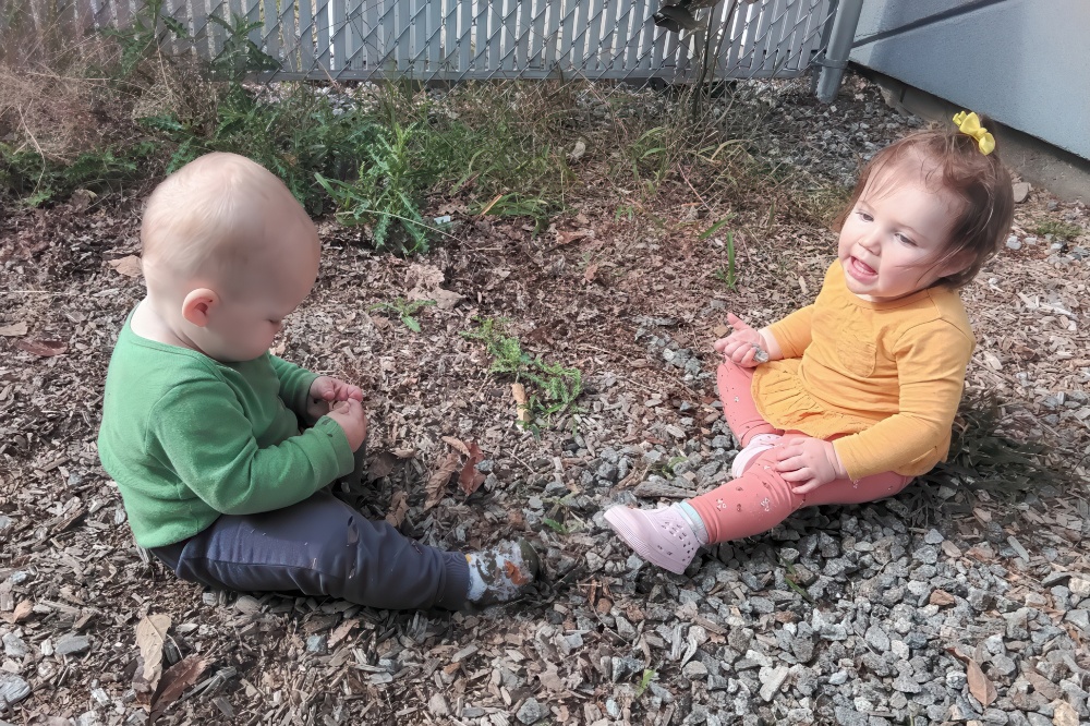 Two playful toddlers sit on the ground surrounded by pebbles and grass, happily engaging with each other.