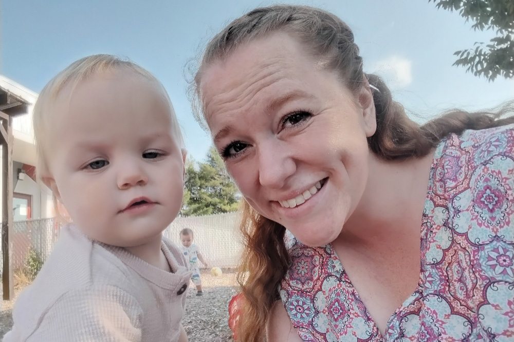 A smiling mother and a toddler pose for a selfie outdoors, with a playful background of other children enjoying the day.