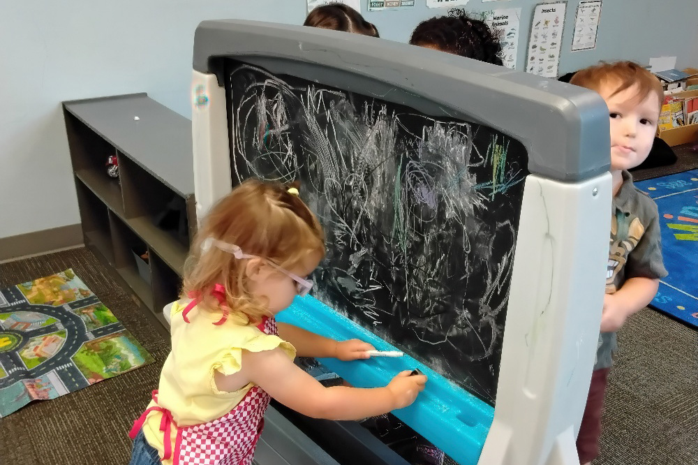 A toddler in a yellow shirt draws on a chalkboard while another child peeks from behind in a classroom.