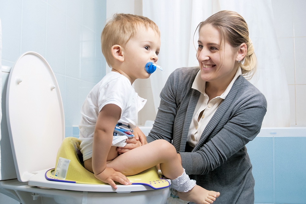 A young child sits on a potty with a pacifier, while a smiling parent offers support and encouragement.