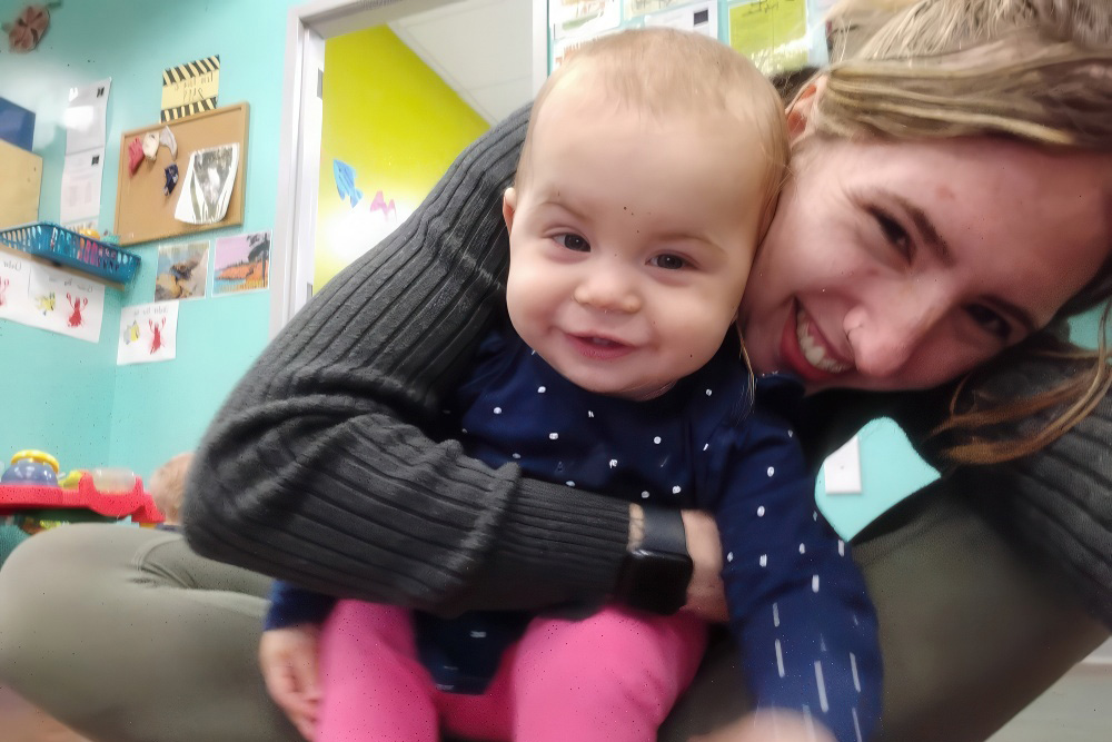 A smiling baby and a teacher share a joyful moment together, both beaming at the camera in a playful indoor setting.