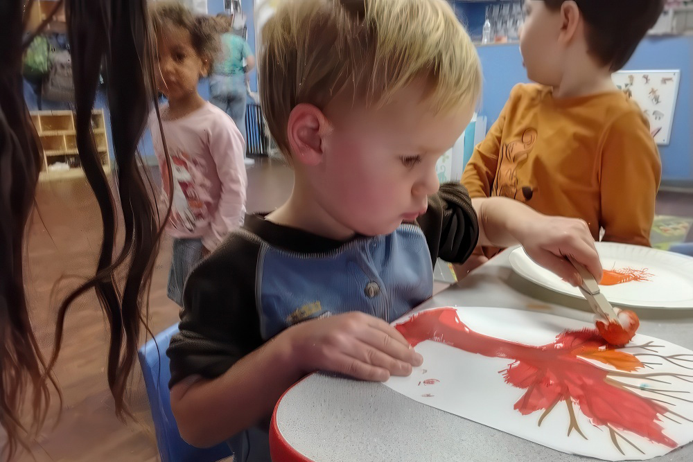 A young boy paints a red tree on paper, focused on his artwork, while other children are engaged in activities nearby.