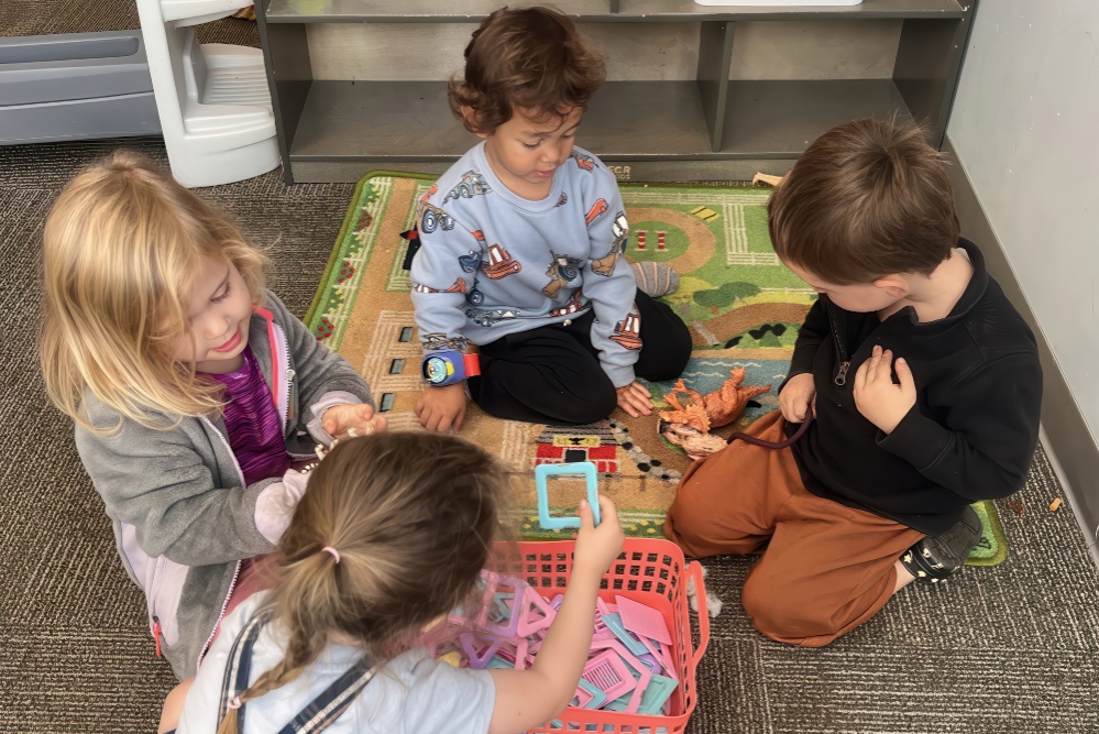 Four children play together on a mat, engaged with toys and colorful shapes, creating a fun and interactive scene.