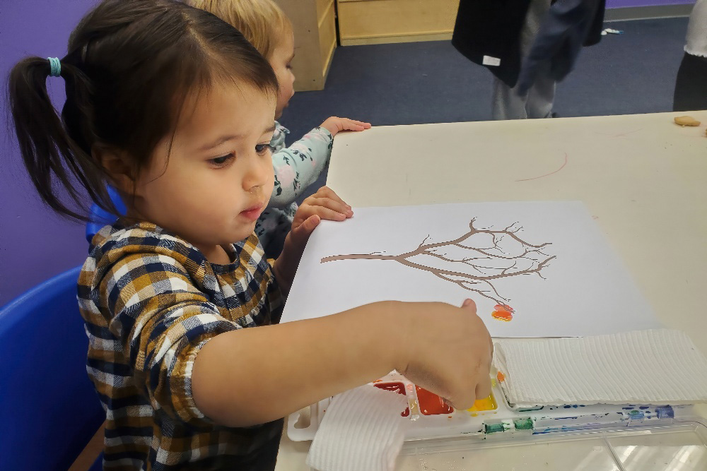 A young girl carefully paints a tree on paper, focused on her artwork at a table with art supplies nearby.