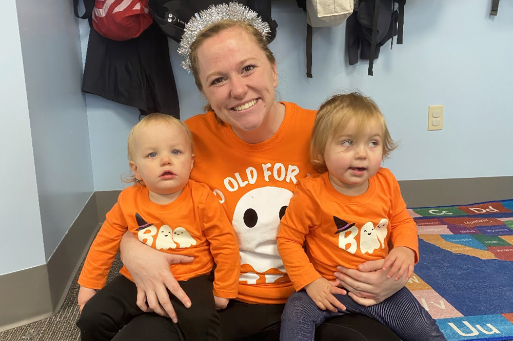 A smiling teacher in an orange shirt sits with two toddlers, dressed in matching outfits, celebrating a festive occasion.