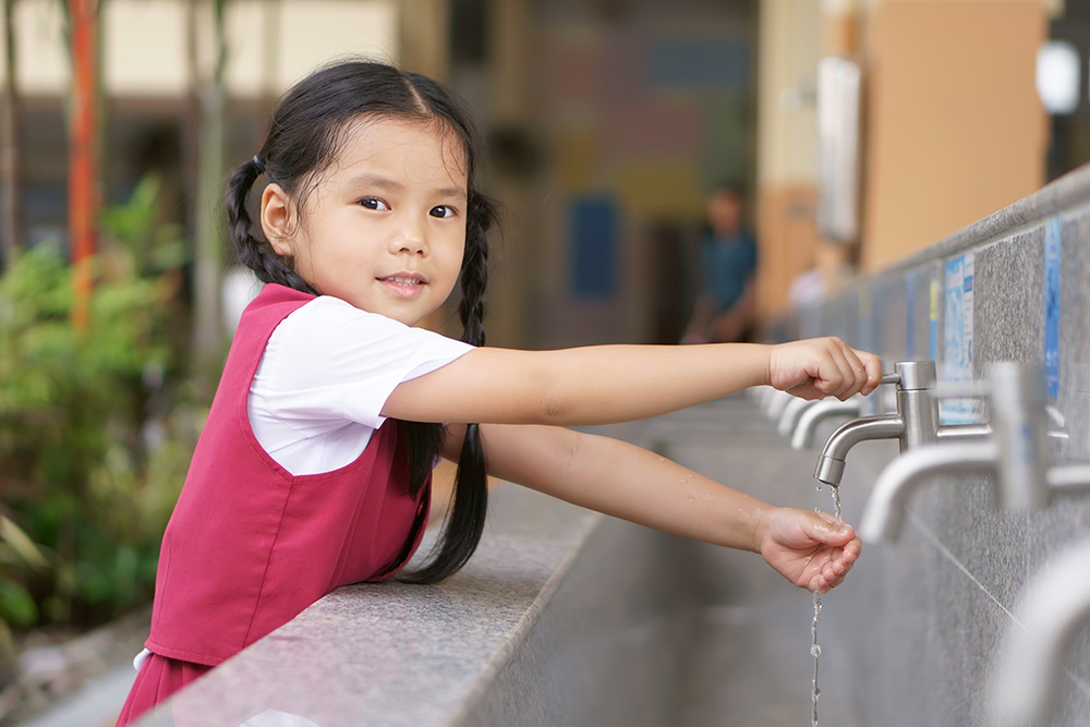 A young girl smiles while washing hands with a water faucet, enjoying a moment of fun outdoors.