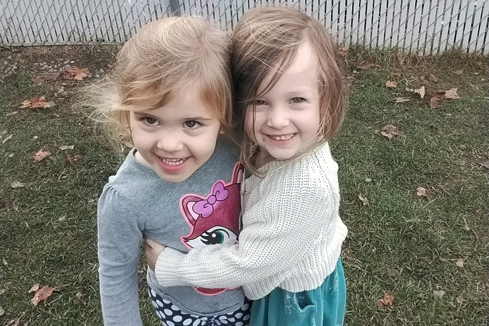 Two girls embrace and smile widely at the camera, radiating joy as they pose together outdoors.