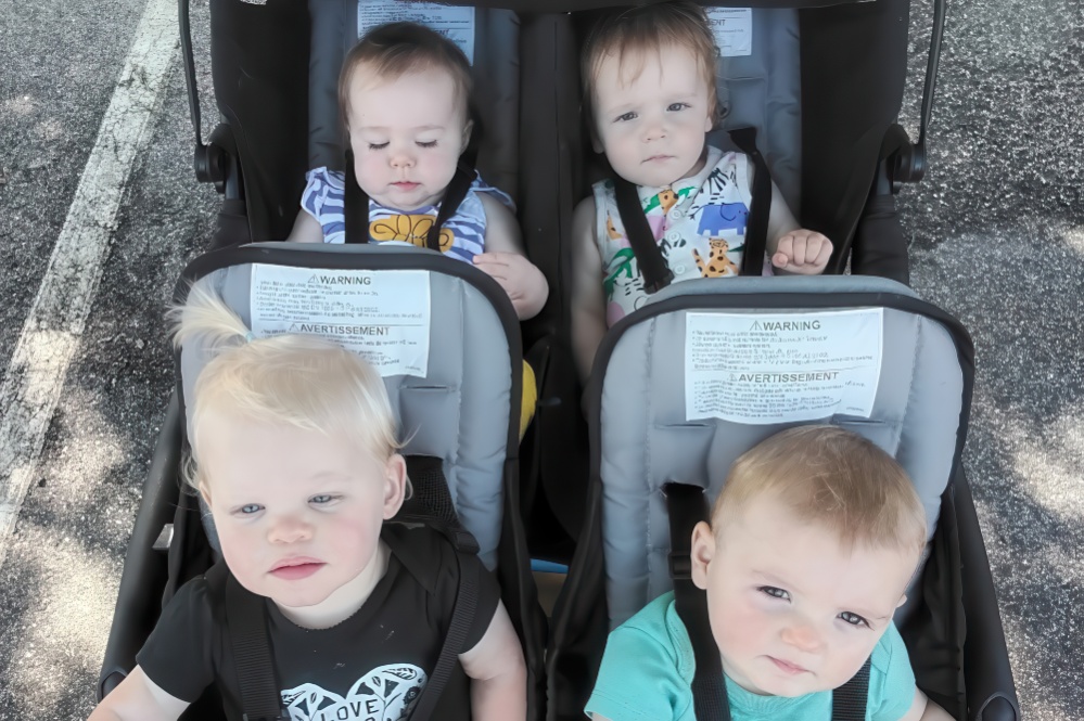 Four toddlers sit in stroller seats, looking curiously at the camera, with bright expressions and playful attire.