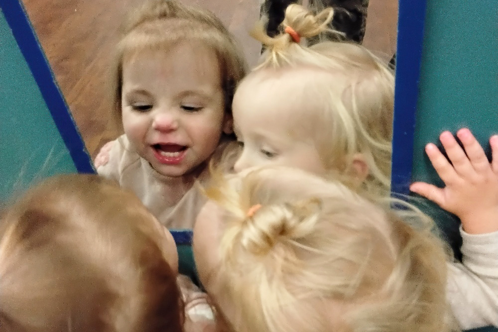 A toddler interacts with their reflection in a mirror, smiling and making funny faces,