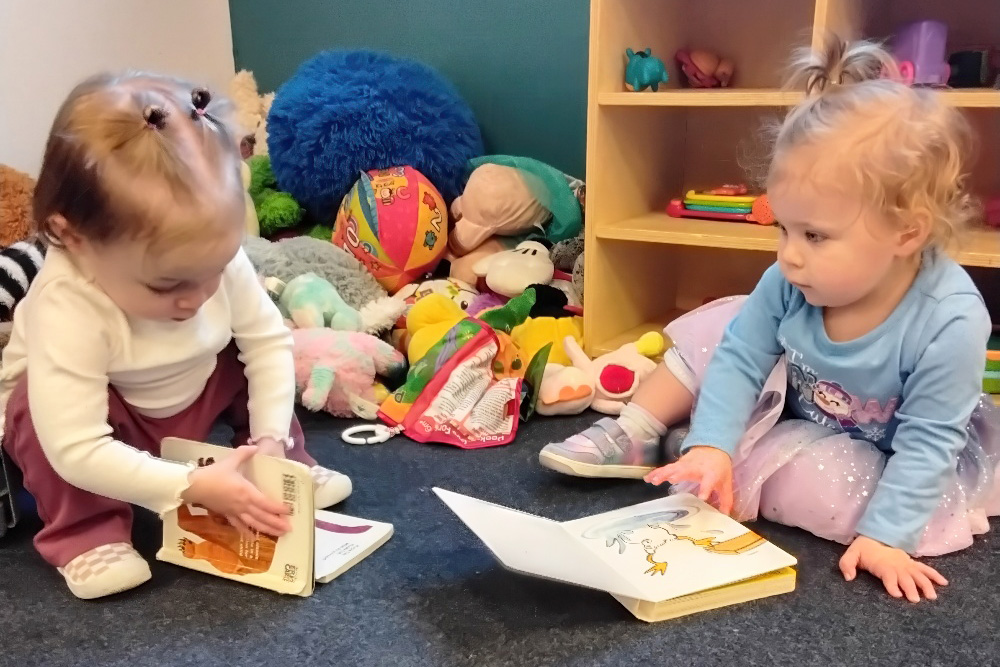 Two toddlers sit on the floor, engrossed in their books amidst a colorful array of toys, sharing a moments of joy.