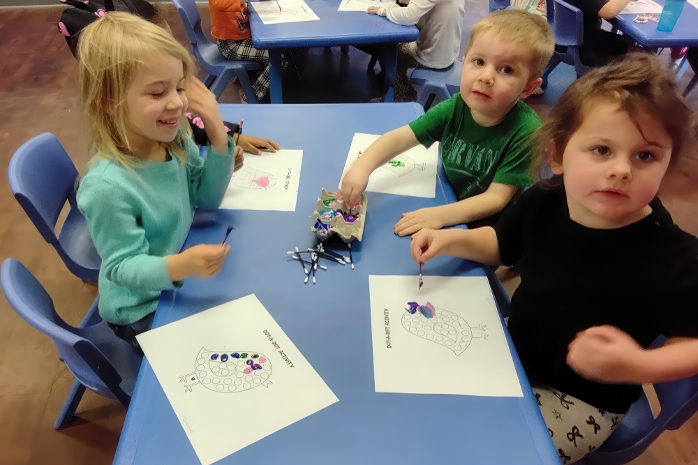 Three children are seated at a table, happily painting colorful pictures, with art supplies scattered around them.