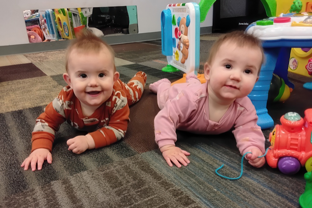 Two happy babies crawl on the floor, playing with toys and smiling at the camera.