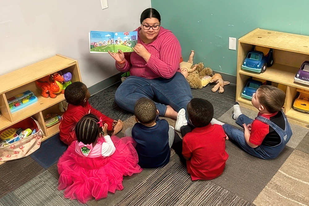 A teacher reads to a group of attentive children sitting on the floor, surrounded by toys in a cozy classroom setting.