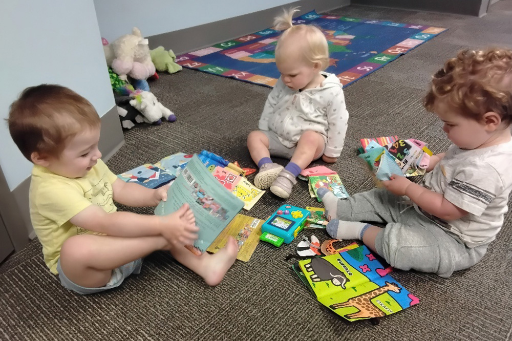 Three toddlers sit on the floor, happily exploring colorful books and toys spread around them.