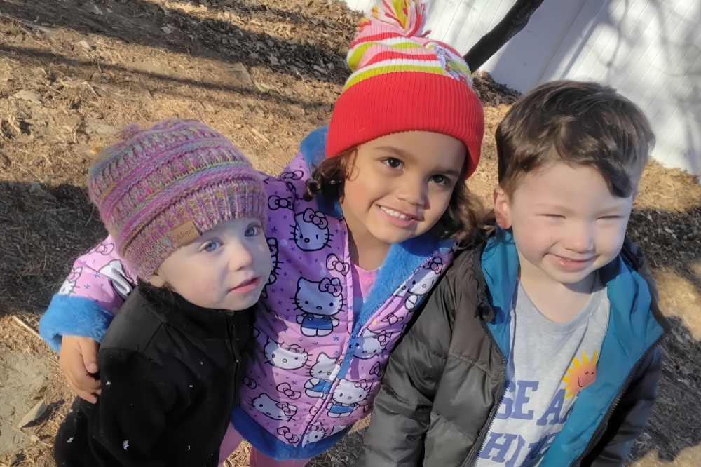 Three children pose together outdoors, wearing colorful hats and jackets, smiling brightly at the camera.