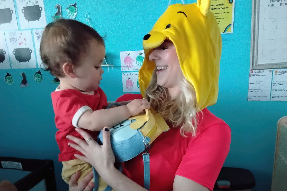 A teacher wearing a Winnie the Pooh hat playfully holds a toddler, both smiling and enjoying a fun moment together.