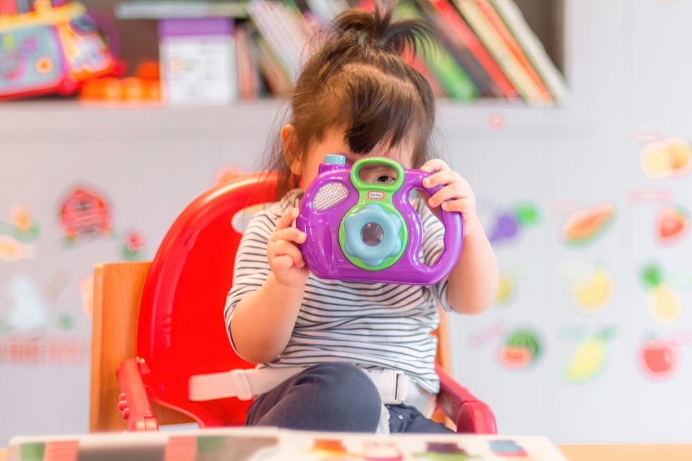 A young child sits confidently, aiming a colorful toy camera at the viewer, ready to capture the moment with delight.