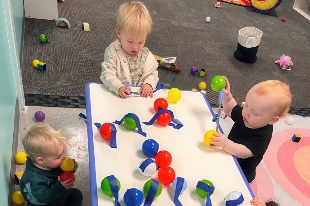 Three toddlers play at a table, exploring colorful balls and ribbons, immersed in creative play in a bright indoor space.