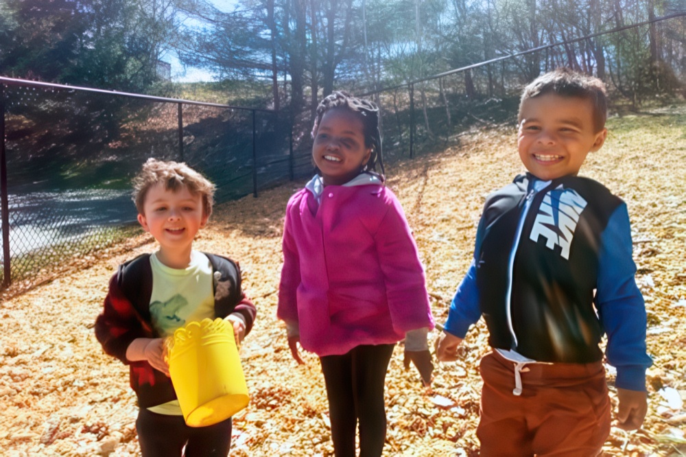 Three children stand together outdoors, smiling brightly among fallen leaves. One holds a yellow bucket.