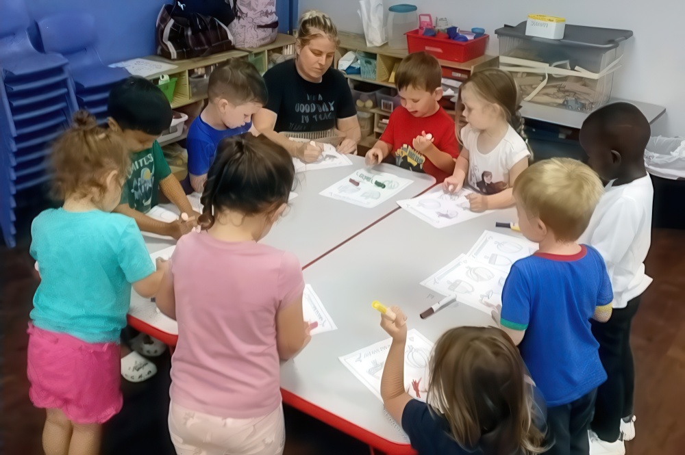 A group of children and a teacher gather around a table, engaged in coloring and crafting, showcasing creativity.