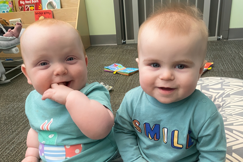 Two cheerful babies sit closely together, both smiling at the camera while wearing matching light blue shirts.