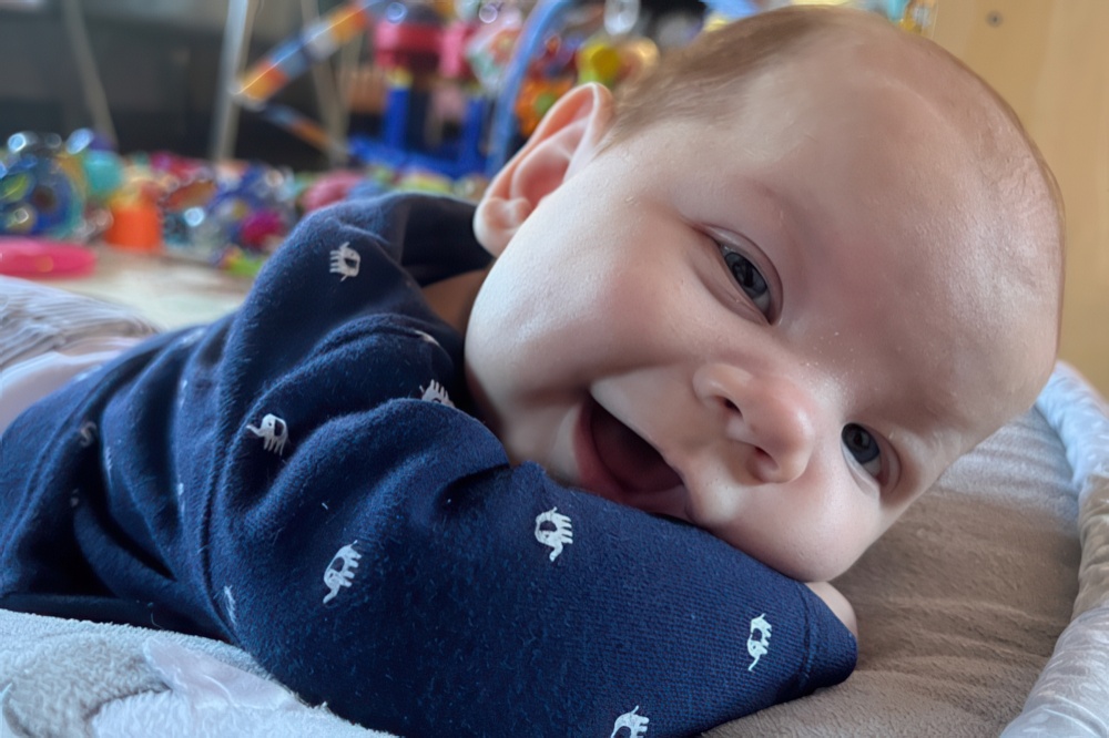 A joyful baby lies on their stomach, smiling brightly at the camera while surrounded by colorful toys.