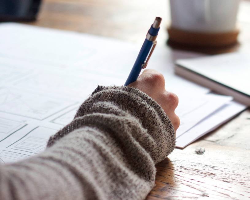 A close-up of a hand holding a pen, focused on writing on paper, with a cozy workspace in the background.